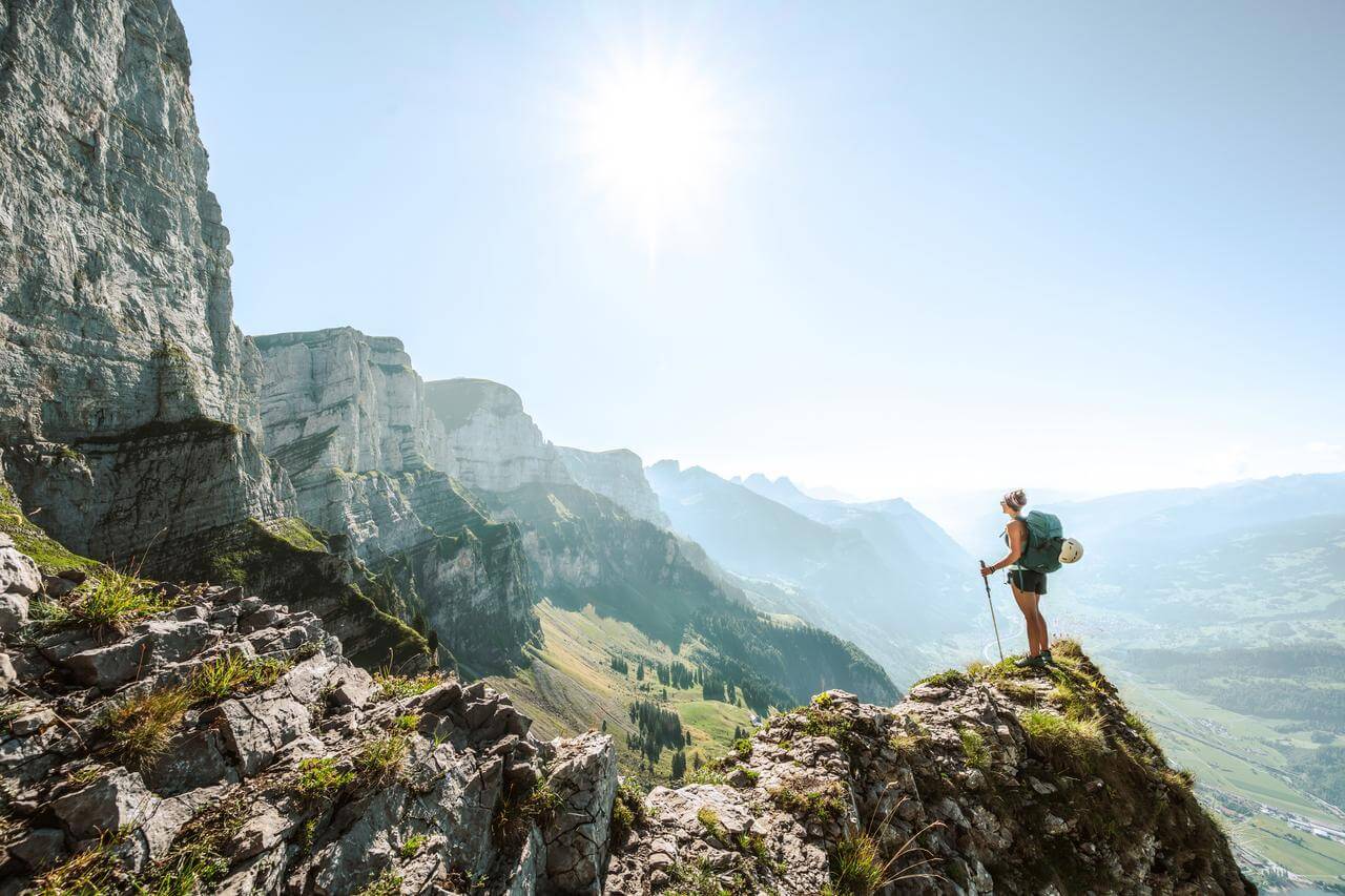 Multi-Risk-Police Eine Person steht auf einem felsigen Berggipfel und blickt in die Ferne. Der Himmel ist klar und die Sonne scheint hell. Im Hintergrund sind steile Bergwände und ein weites Tal sichtbar.