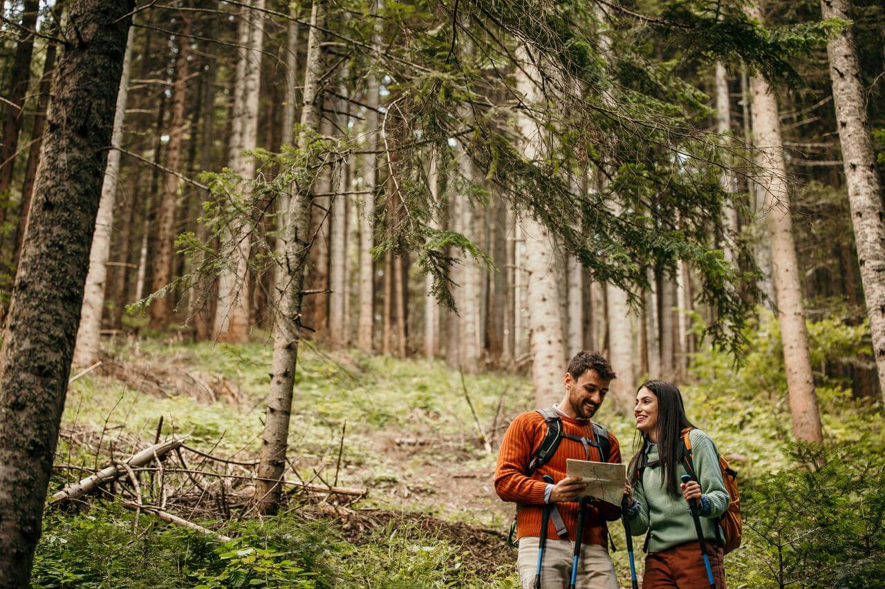 Multi-Risk-Police Ein Mann und eine Frau wandern im Wald und betrachten eine Karte. Beide tragen Rucksäcke und Outdoor-Kleidung. Die Umgebung ist voller Bäume und grünem Laub.