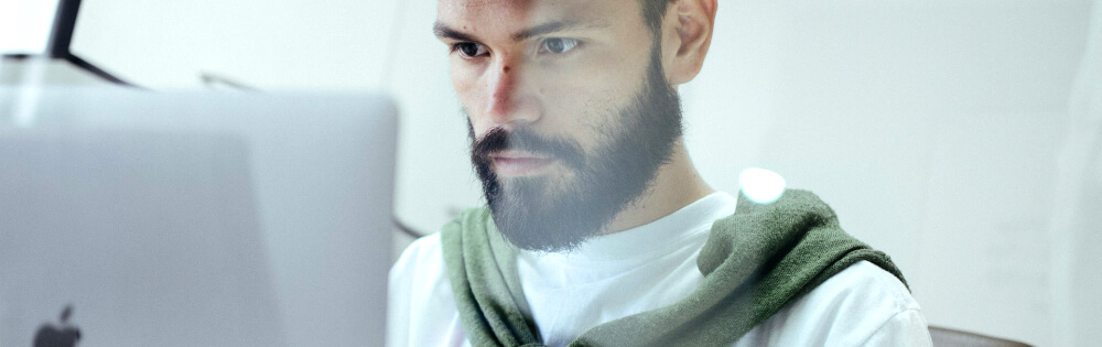 Cyberversicherung Ein Mann mit Bart und ernstem Gesichtsausdruck, der auf einen Laptop schaut. Er trägt ein weißes T-Shirt und hat einen grünen Pullover um die Schultern gelegt.