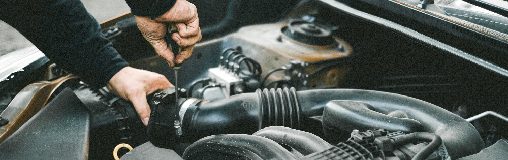 Kfz-Werkstätten-Police Ein Mechaniker repariert den Motor eines Autos, indem er mit Werkzeug einige Teile anpasst. Im Bild ist ein offener Motorraum mit verschiedenen Autoteilen zu sehen.