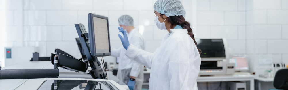 Elektronikversicherung Zwei Laborfachkräfte tragen Schutzkleidung, einschließlich Laborkittel, Haarnetz und medizinischer Masken, während sie an einem Computerbildschirm arbeiten. Sie befinden sich in einem modernen Labor mit verschiedenen Geräten.